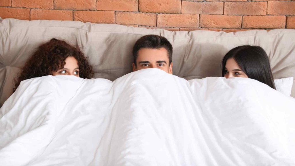 Image of a man and two women in bed under the covers in a threesome.